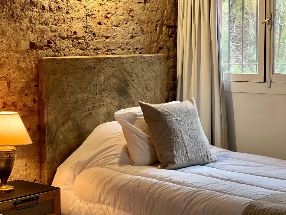 A bed made up against an exposed stone wall at Estancia La Loma, with a lit lamp and a window onto the park